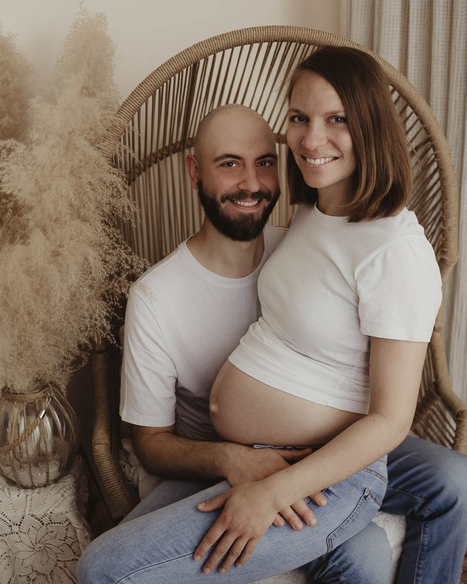 Babybauchfotografie: Ein Paar im bequemen Korbstuhl, die Mama zeigt stolz den Bauch.